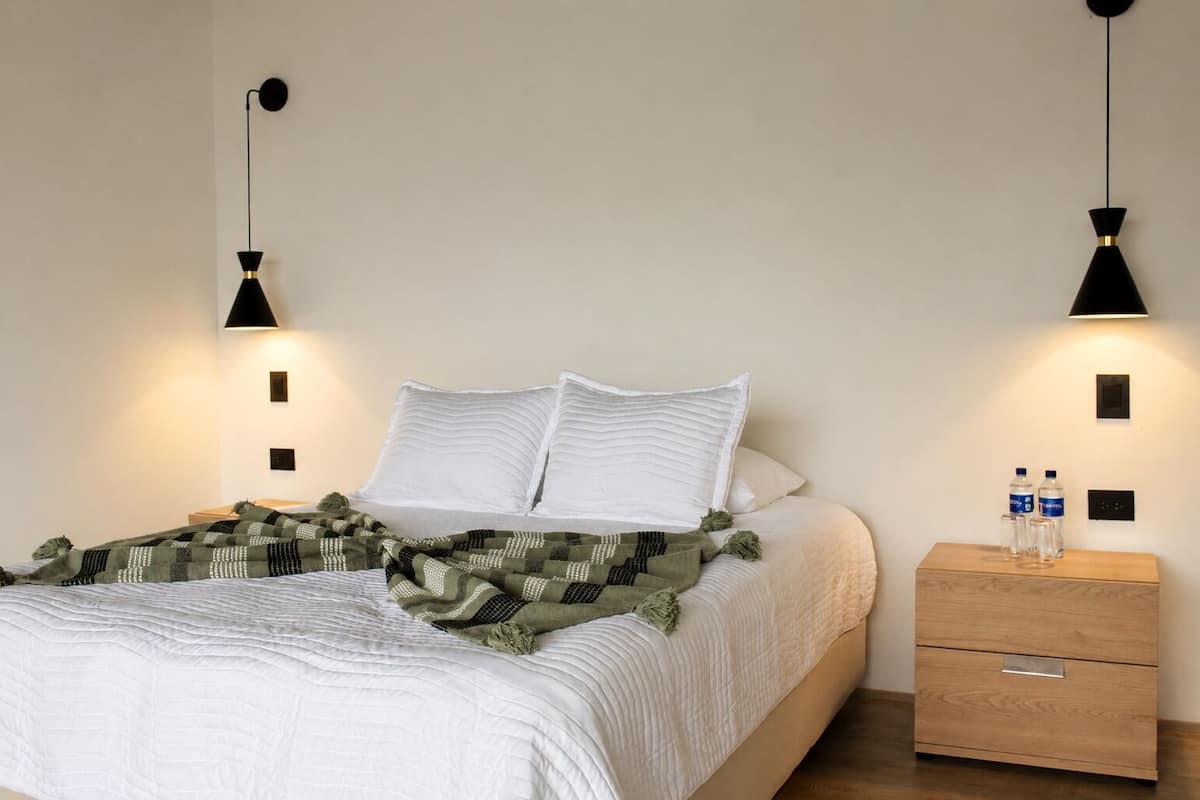 A neatly made bed with white bedding, a green and black patterned throw, and two black wall-mounted lamps.