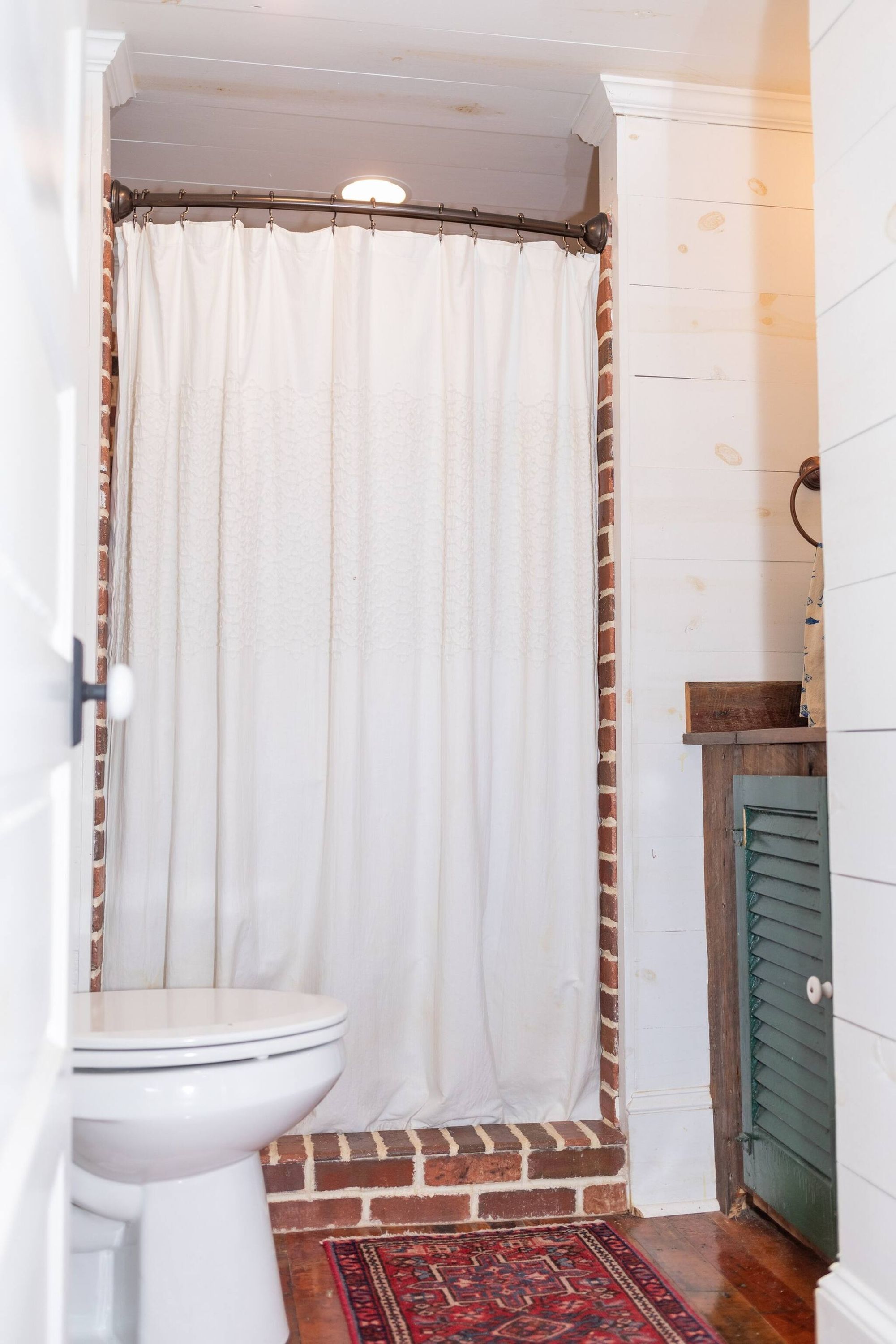 A bathroom with a white toilet, a shower curtain, a wooden bench, and a red patterned rug.