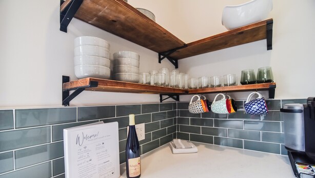 A kitchen with wooden shelves holding plates, glasses, and a wine bottle. There is a menu stand and a coffee machine on the counter. - Charming Downtown Fort Mill Home w/ Large Backyard (Fort Mill)