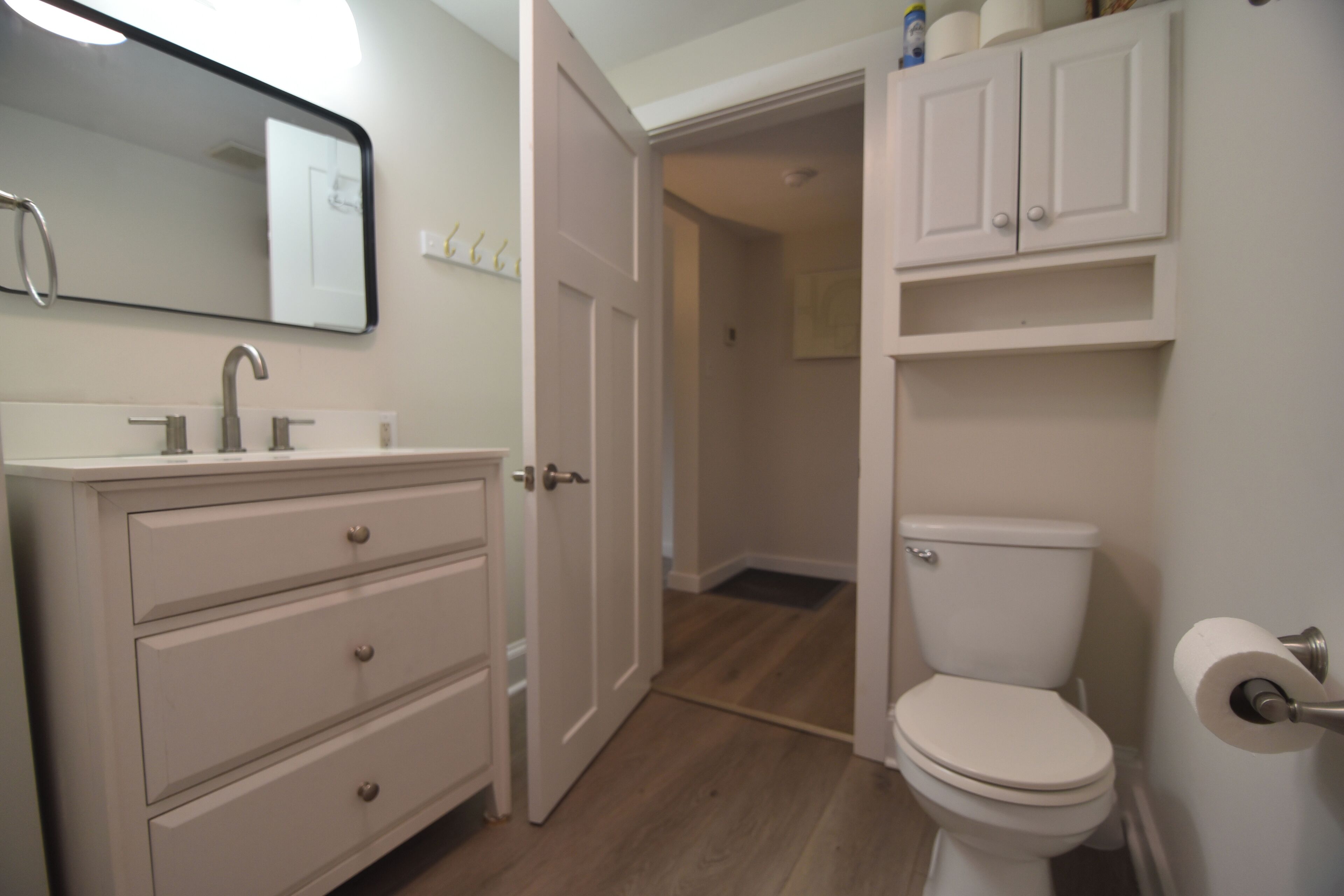 A bathroom with a white sink, mirror, and toilet. There is a cabinet above the toilet and a door leading to another room.
