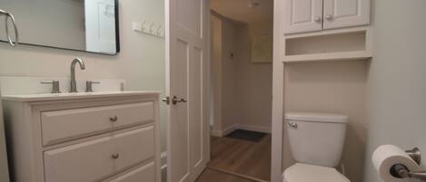 A bathroom with a white sink, mirror, and toilet. There is a cabinet above the toilet and a door leading to another room.