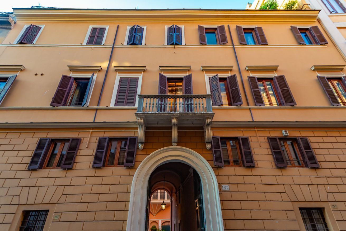 A multi-story building with a central entrance, multiple windows with shutters, and a balcony with a railing.