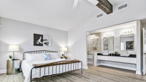 Une chambre à coucher comprenant un lit, des lampes de chevet, un miroir fixé au mur, et une salle de bain avec un meuble-lavabo et un miroir.