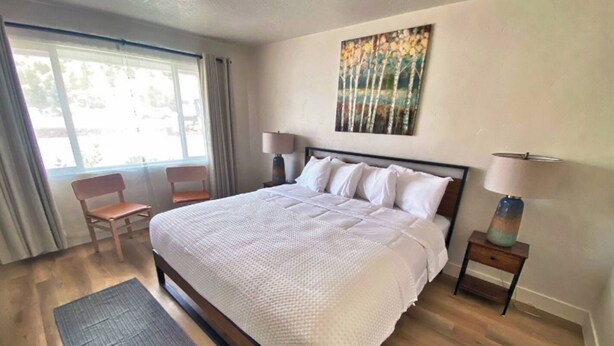 Een netjes ingerichte slaapkamer met een groot bed, twee nachtkastjes met lampen, een stoel en een raam met gordijnen.