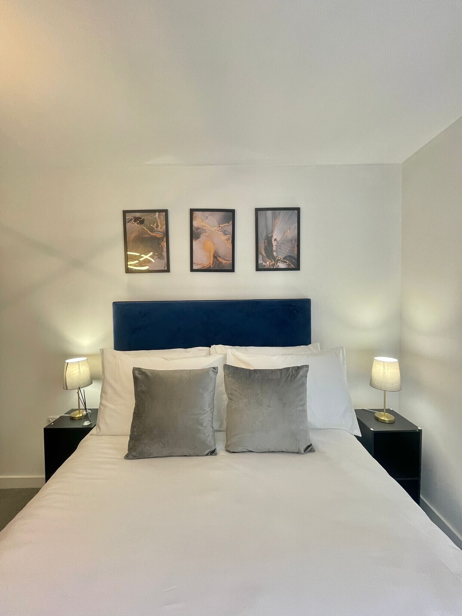 A neatly made bed with white linens, two gray pillows, and a blue headboard. There are three framed artworks on the wall above the bed. Two bedside tables with lamps are on either side of the bed.