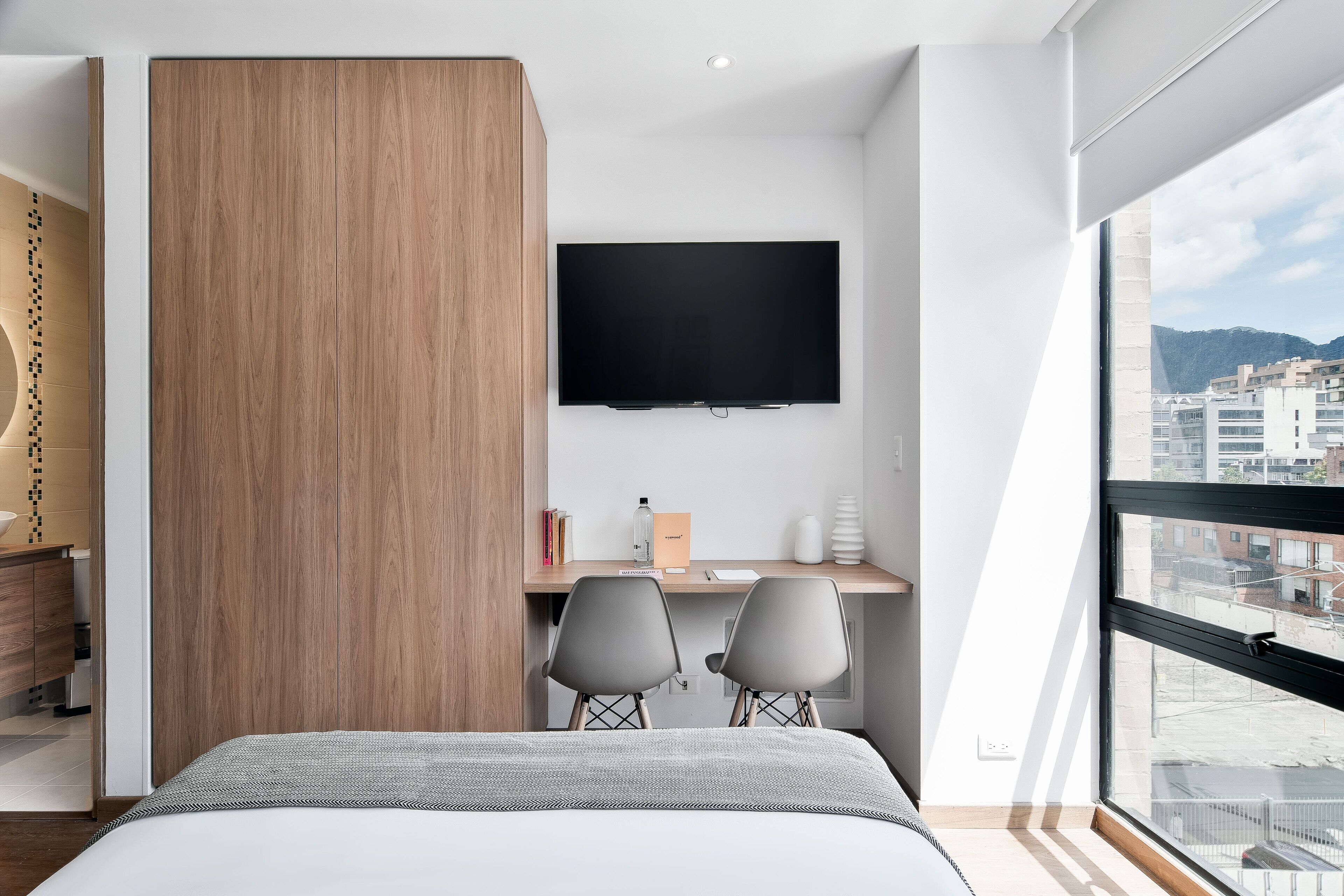 A modern hotel room with a large bed, a desk with two chairs, a flat-screen TV, and a large window offering a city view.