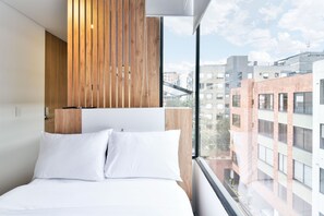 A modern hotel room with a large bed, wooden panel wall, and a view of the cityscape. - Nuevetres by Wynwood House (Bogotá)
