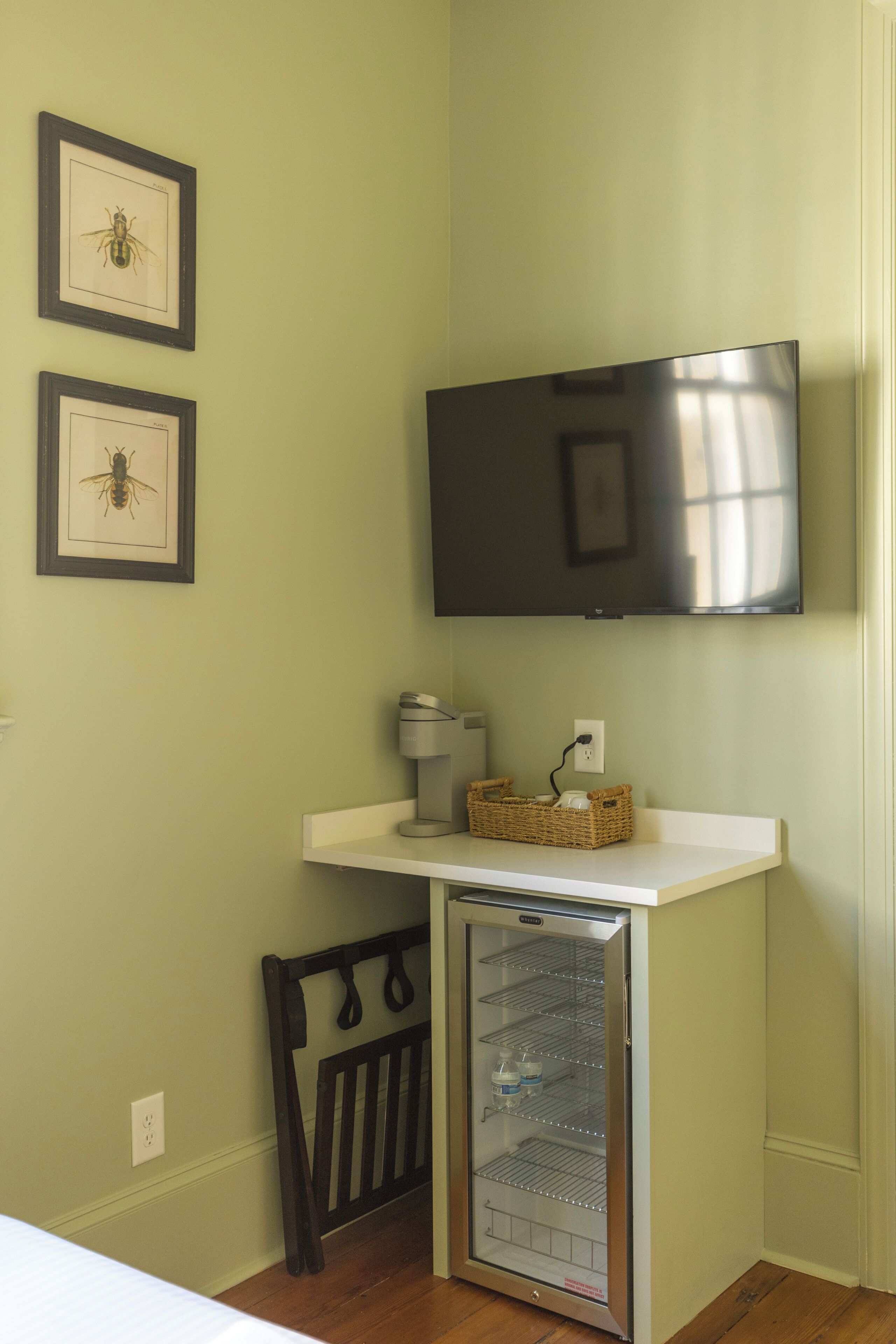 Una pequeña kitchenette con refrigerador, microondas y lavamanos. Hay cuadros en la pared y un televisor de pantalla plana montado sobre la encimera.