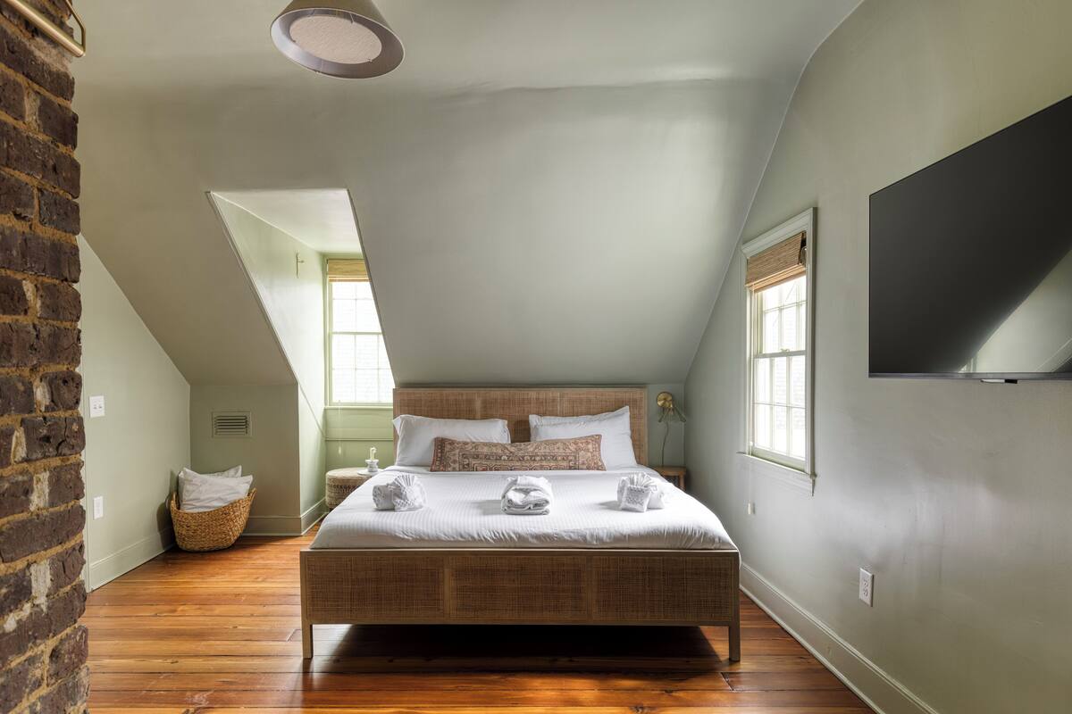 A bedroom with a large bed, a TV mounted on the wall, and a window with blinds.