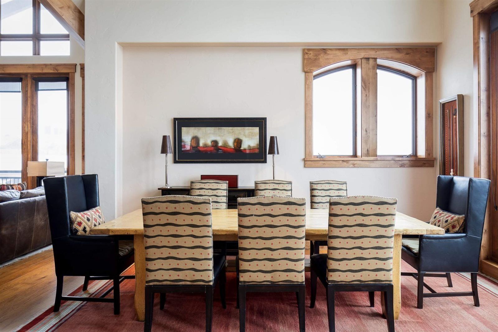 Een eetruimte met een houten tafel en zes gestreepte stoelen, een kunstwerk in een lijst erboven, en grote ramen met houten kozijnen.