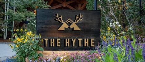 A wooden sign with a logo and the text 'THE HYTHE' in front of a building with a sloped roof and wooden beams.