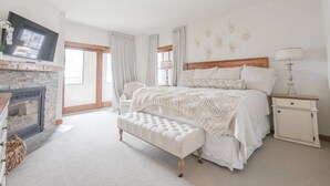 A bedroom with a large bed, a bench, a nightstand, a lamp, a fireplace, and a TV mounted on the wall.