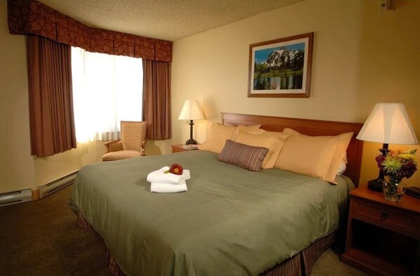 Una habitación de hotel con una cama grande, dos mesitas de noche con lámparas, una silla, un cuadro en la pared y un ventanal con cortinas.