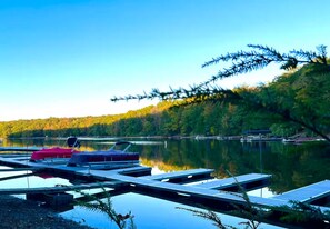 A lakeside dock with boats, a covered area, and a forested shoreline. - Rustic Charm Meets Modern Boutique + Lake Central + Hot Tub, Sauna, Lake Access (Oakland)
