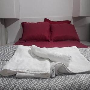 En säng med ett geometriskt mönstrat överkast, två röda kuddar och en vikt vit handduk på sängen.