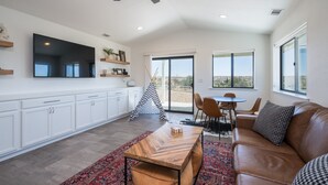 A spacious living area with a large flat-screen TV, a ceiling fan, a dining table with chairs, a sofa with patterned cushions, and a large window offering a view of the outdoors. - Wine Country Escape*Hot Tub*Fire Pit (Paso Robles)