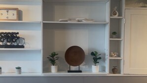 A room with a grey wall, a white shelf with a clock and potted plants, and a door with a decorative item. - Modern Kentucky Retreat Near Ark & Cincinnati! (Covington)