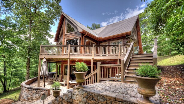 A two-story log cabin with a balcony, stone steps, and potted plants.