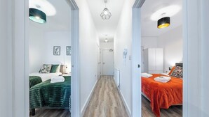 Hotellihuone, jossa on puulattia, kaksi sänkyä vihreillä ja oransseilla vuodevaatteilla, käytävä, jossa on kattovalaisin, ja ovi käytävän päässä.