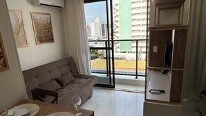 A modern living room with a sofa, dining table set for two, and a large window with a city view. - Melhores Flats - Belezinha perto do mar (João Pessoa)