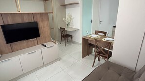 A modern living room with a flat-screen TV, a sofa, a dining area with a table and chairs, and a kitchen area with cabinets and a countertop. - Melhores Flats - Belezinha perto do mar (João Pessoa)