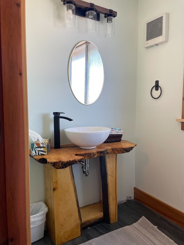Baño con tocador de madera, lavabo blanco y espejo redondo. Hay un perchero y un cubo de basura pequeño.