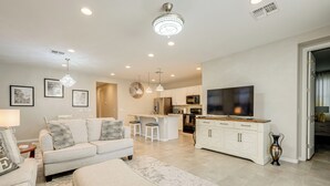A modern living room with a sofa, ottoman, and a TV. There is a kitchen area with a refrigerator, microwave, and island. - Central Casa Grande Home in Quiet Neighborhood (Casa Grande)