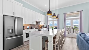 A modern kitchen with white cabinetry, a stainless steel refrigerator, a marble island, pendant lighting, and a view of the outdoors through a large window.