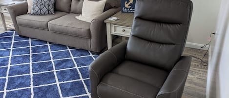 A living room with a brown leather chair, a brown sofa with patterned cushions, a blue and white rug, a small table with a lamp, and a decorative plant.