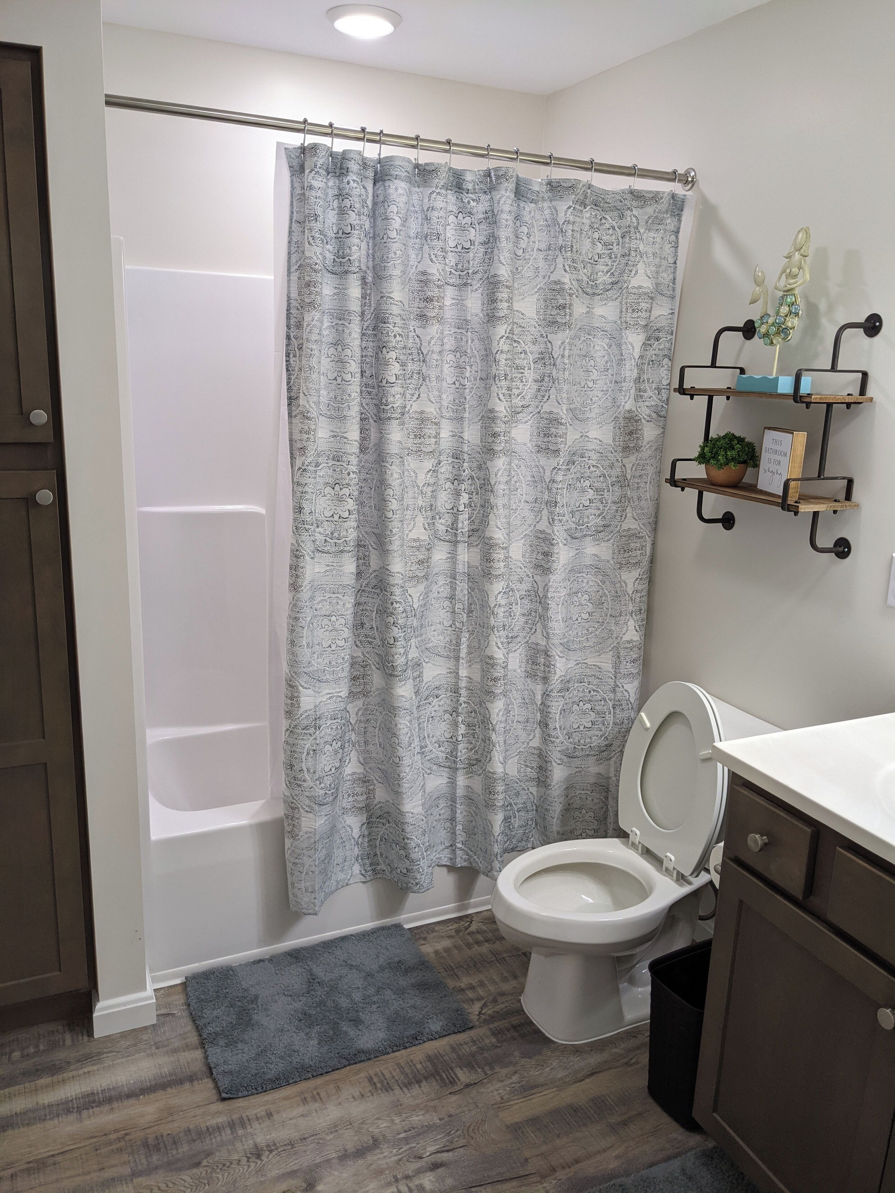 A bathroom with a shower curtain, a toilet, a small shelf with a plant, and a grey rug.
