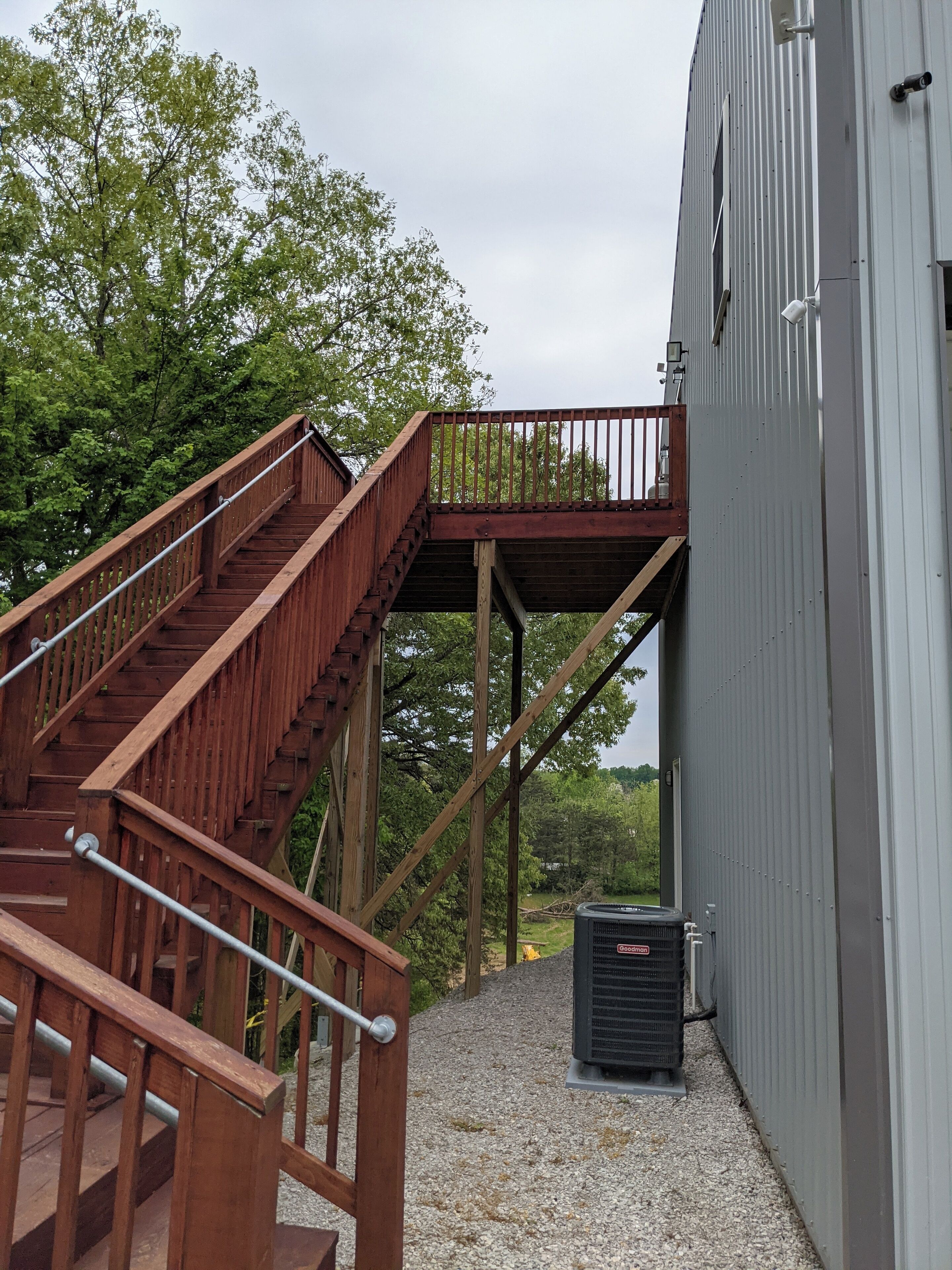 A wooden staircase leading to a balcony, a grey building, and an air conditioning unit on a gravel surface.