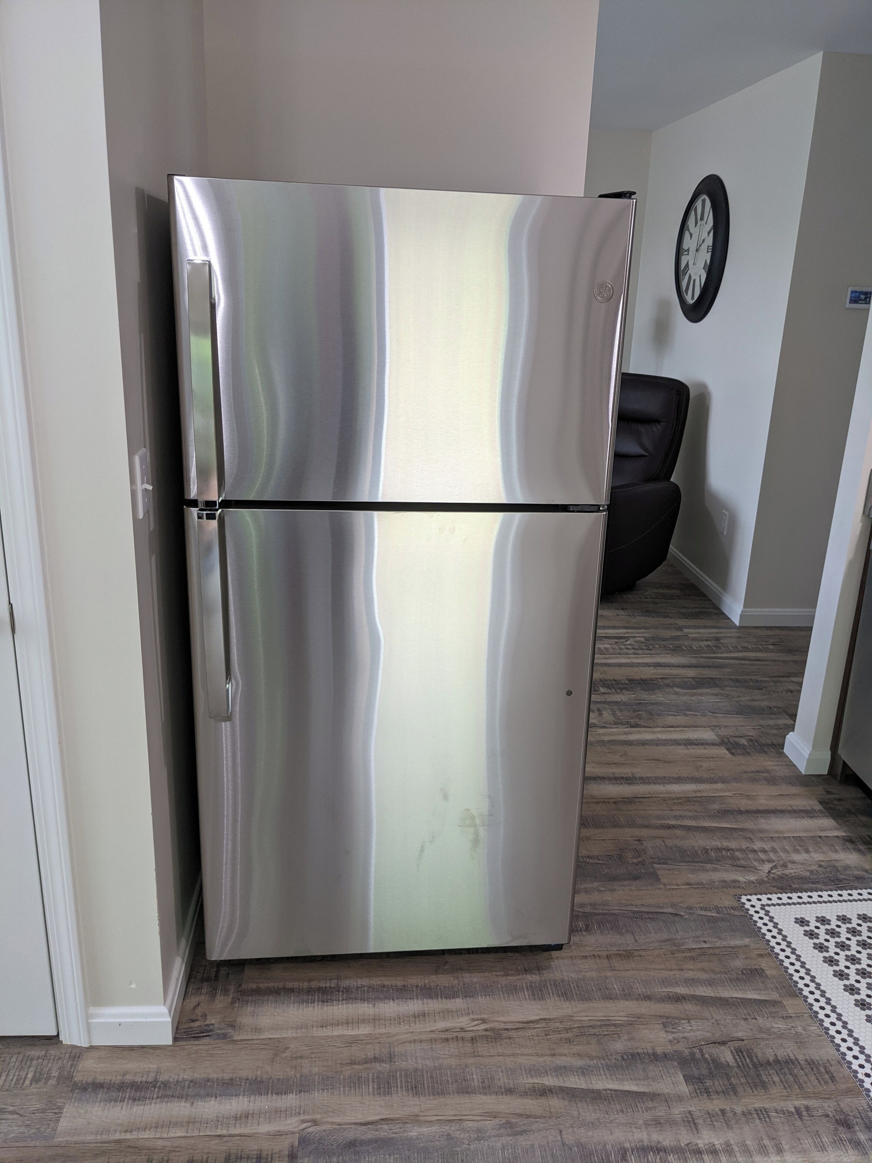 A stainless steel refrigerator in a kitchen with wooden flooring and a clock on the wall.