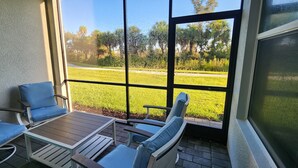 A balcony with two blue cushioned chairs and a small table, overlooking a well-maintained lawn and trees.