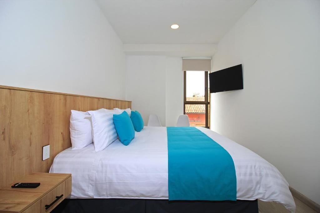 Camera d'albergo con una testiera in legno, un letto con biancheria da letto bianco e blu, una TV a parete e una finestra con vista sugli edifici.