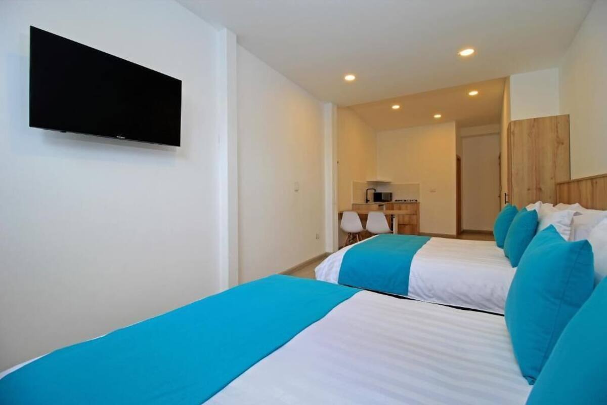 A hotel room with two beds, a flat-screen TV, a kitchenette, and a bathroom visible in the background.