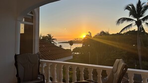 A balcony with a view of a sunset over water, palm trees, and a tiled roof. - Oceanfront Penthouse in Puerto Plata (Puerto Plata)