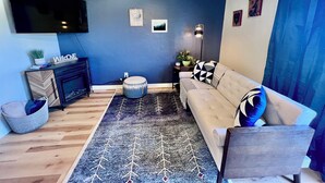 Un soggiorno moderno con una parete azzurra a contrasto, una TV a schermo piatto sopra un caminetto, un divano con cuscini decorati e un poggiapiedi rotondo.