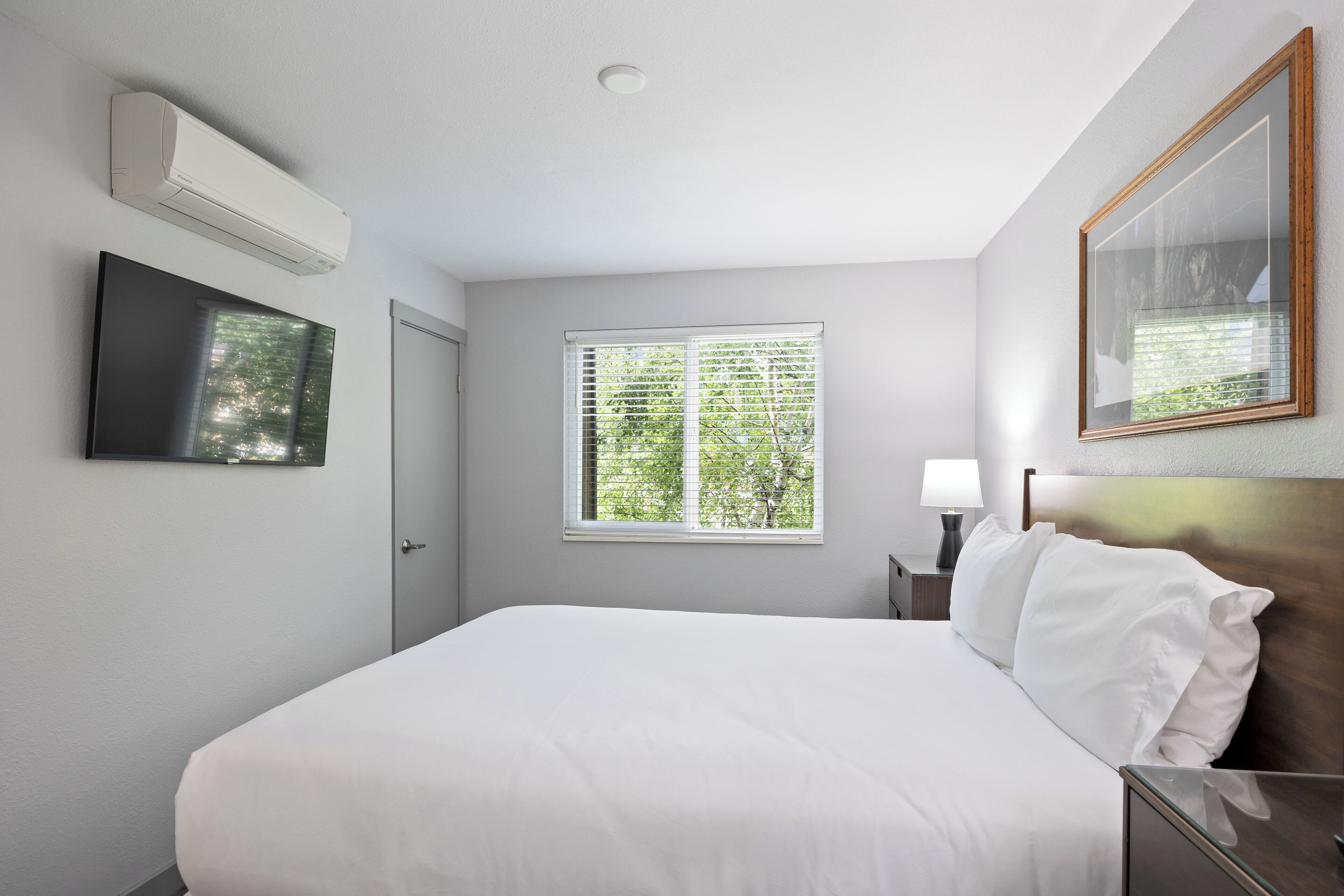 A hotel room with a bed, a TV, a window with blinds, a bedside lamp, and an air conditioning unit.
