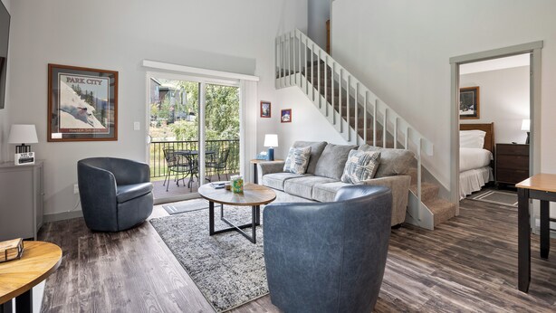 Una sala de estar moderna con un sofá gris, un sillón azul, una mesa de centro de madera y una escalera que conduce a un nivel superior.