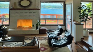 A living room with a fireplace, leather chairs, a sofa, and large windows overlooking a body of water.