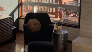 A room with a large window overlooking a lake and forest, a black chair with a cushion, a small round table with a candle, and a decorative vase.