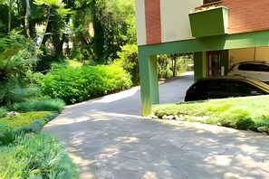 A paved driveway leading to a building with a green and red facade, surrounded by lush greenery and a parked car.