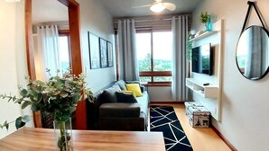 Un soggiorno moderno con un tavolo da pranzo in legno, un divano con cuscini, una TV su un mobile e un'ampia finestra con tende.