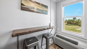 Une pièce avec une étagère en bois, deux tabourets, un tableau et une fenêtre donnant sur un paysage verdoyant.