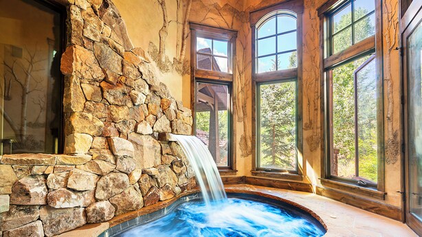 Un jacuzzi de paredes de piedra con cascada, ventanas amplias y vistas a árboles.