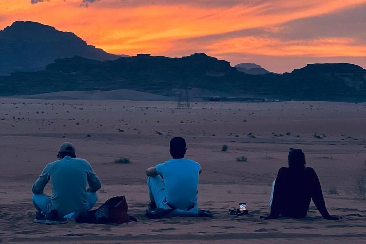 Trois personnes sont assises sur le sable, dans un paysage désertique, avec des montagnes en arrière-plan et un ciel au coucher du soleil.