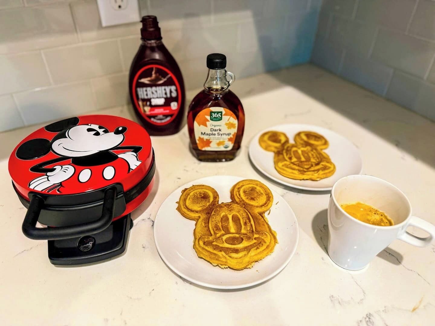 Eine Küchenarbeitsplatte mit einem Mickey-Mouse-Toaster, zwei Tellern mit Mickey-Mouse-Pfannkuchen, einer Tasse sowie Flaschen mit Hershey's-Sirup und Ahornsirup.