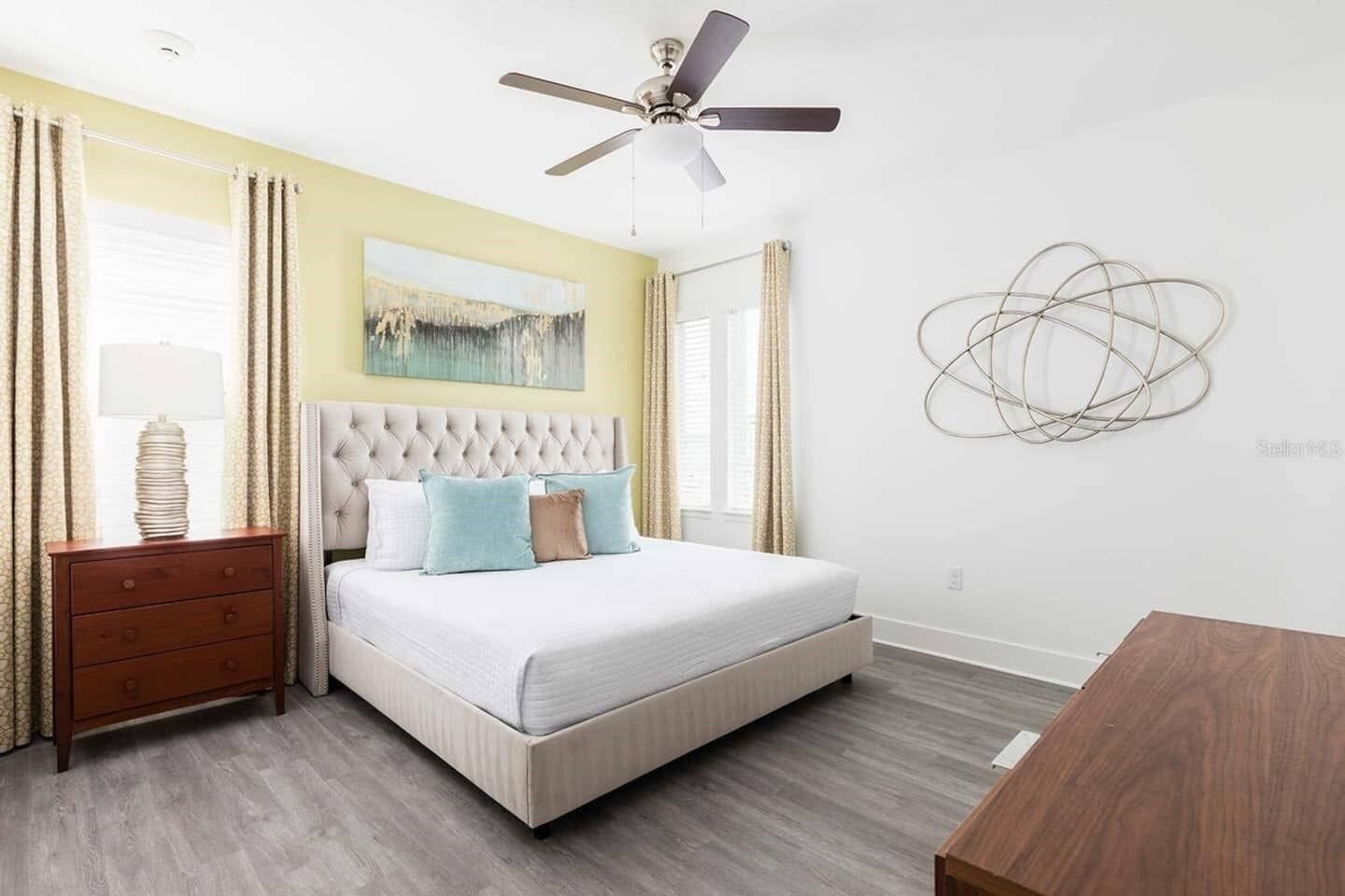 Ein Schlafzimmer mit einem gepolsterten Kopfteil, einem Bett mit weißer Bettwäsche, einem hölzernen Nachttisch, einem Deckenventilator und einem an der Wand befestigten abstrakten Kunstwerk.
