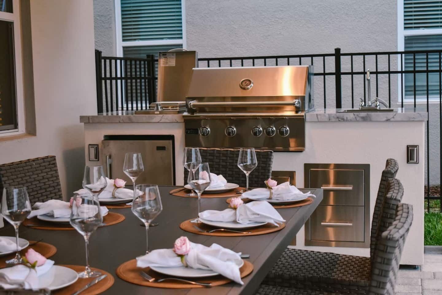 Un espace extérieur aménagé pour les repas, doté d’un barbecue et d’un four intégrés, entouré de chaises et agrémenté de verres à vin et d’assiettes.