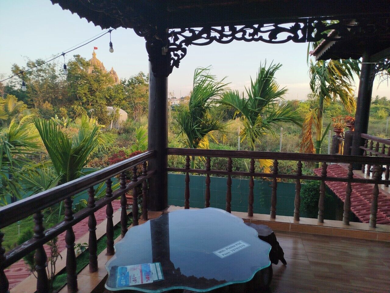 A balcony with a glass table, wooden railing, and a view of greenery and a building in the background.